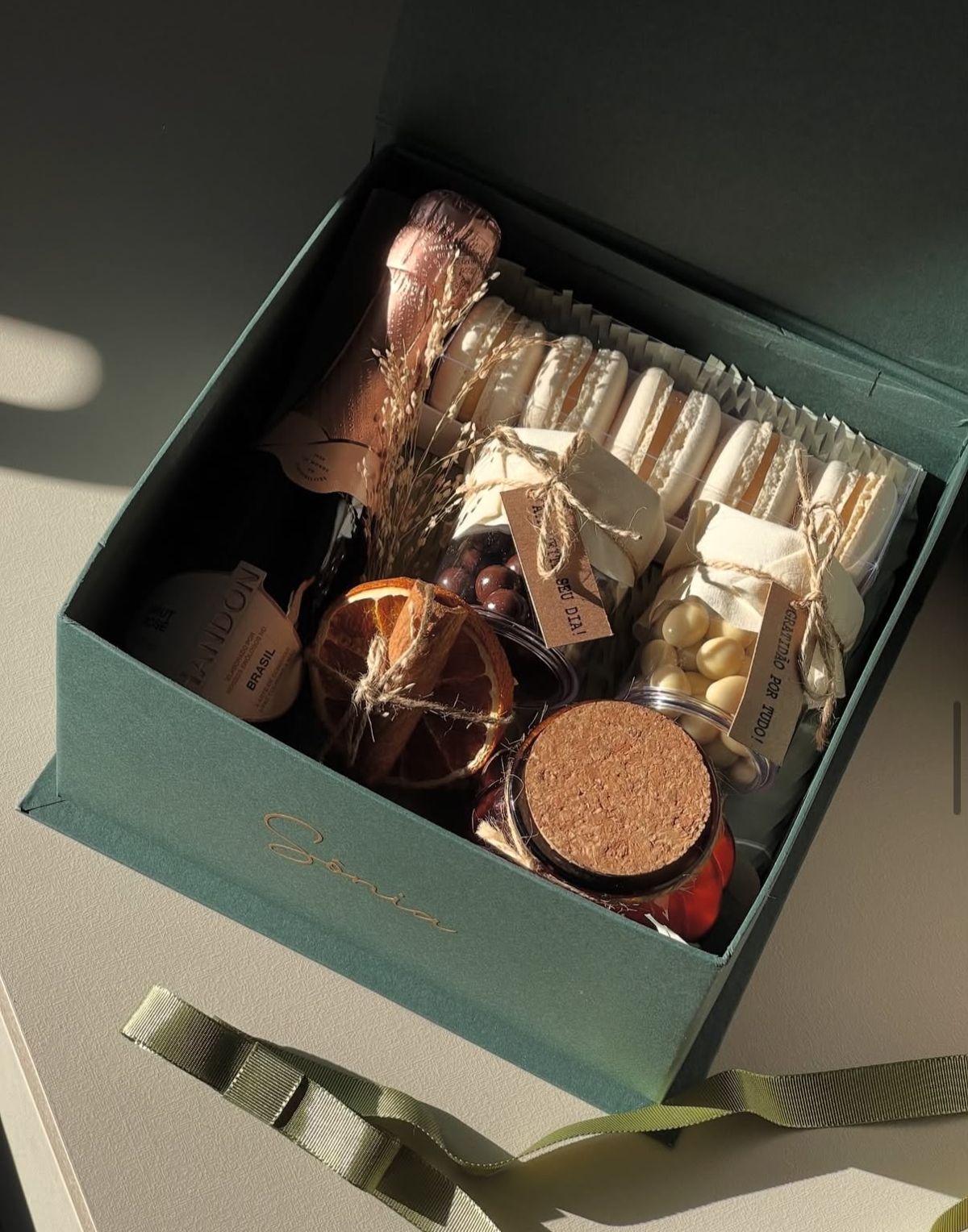 Caixa cartonada verde aberta com espumante, macarons e potes de chocolate